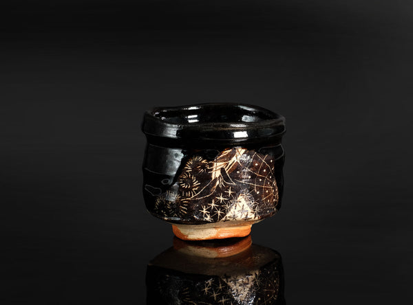 A small glossy black ceramic cup with intricate gold floral and star-like engravings, set against a dark reflective background.