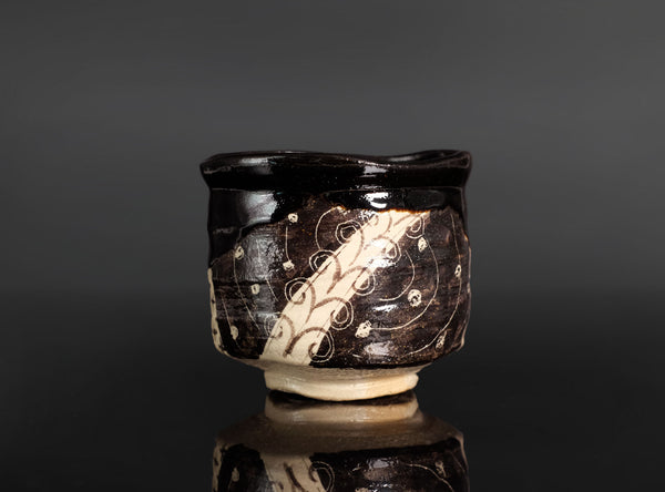 A handmade ceramic tea whisk holder or small bowl with a glossy black glaze and cream-colored abstract swirl patterns, set against a dark gray background with a soft reflection below.