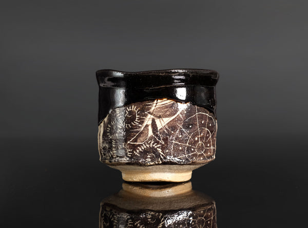 A handmade ceramic tea cup with a glossy black top glaze and a textured, crackled brown and beige body, shown on a reflective dark gray background.