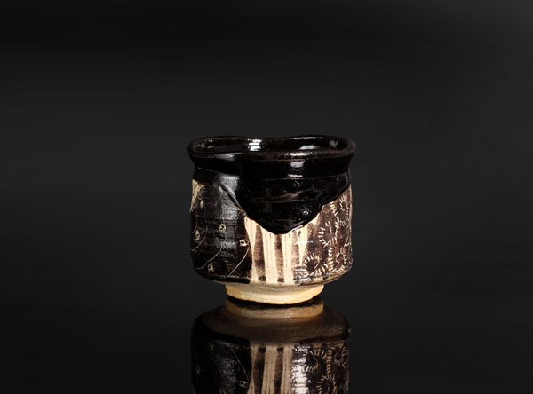 A handmade ceramic cup with a glossy black glaze and hand-painted floral patterns on the sides, set against a dark reflective background.