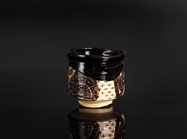 A handmade ceramic tea cup with a glossy black glaze and a textured, patterned brown and beige body, set against a dark reflective background.