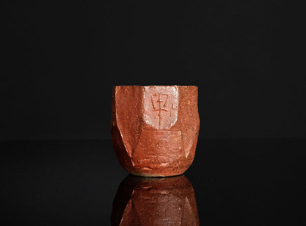 A handmade terracotta ceramic bizen guinomi with a faceted, geometric design and a carved kanji character, displayed on a reflective black surface against a solid black background.