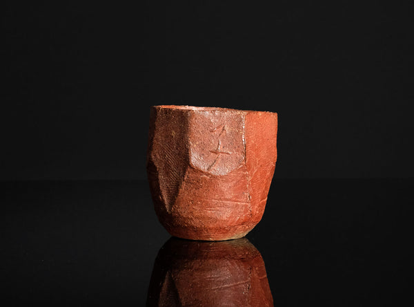 A handmade terracotta ceramic bizen guinomi with a faceted, geometric design and a carved kanji character, displayed on a reflective black surface against a solid black background.