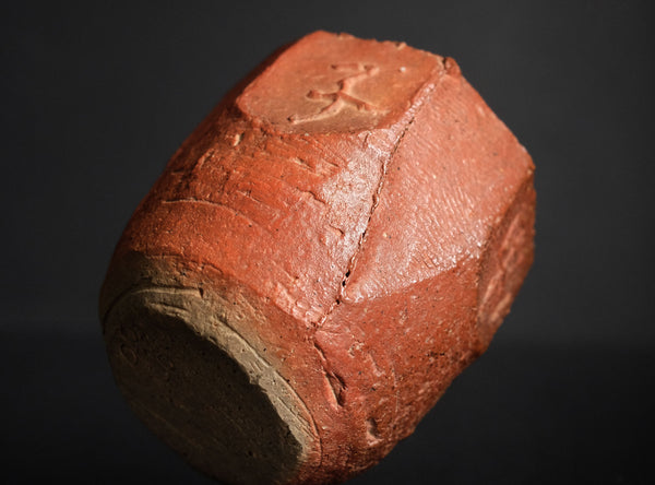 A handmade terracotta ceramic bizen guinomi with a faceted, geometric design and a carved kanji character, displayed on a reflective black surface against a solid black background.