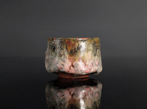 A handmade ceramic sake cup with a textured, organic glaze featuring mottled shades of brown, gray, pink, and gold, set against a dark reflective background.
