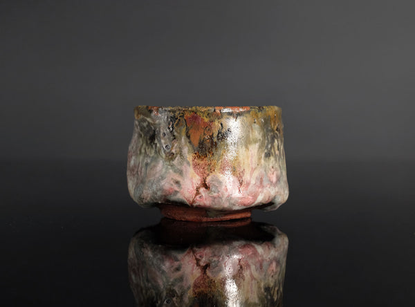 A handmade ceramic sake cup with a textured, organic glaze featuring mottled shades of brown, gray, pink, and gold, set against a dark reflective background.
