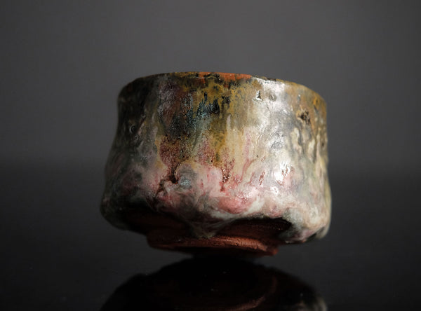 A handmade ceramic sake cup with a textured, organic glaze featuring mottled shades of brown, gray, pink, and gold, set against a dark reflective background.