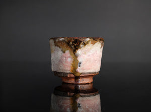 A handmade ceramic sake cup with a textured, organic glaze in pink, brown, and white, featuring a dripping amber glaze detail, set against a dark gray reflective background.