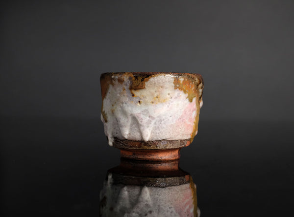 A handmade ceramic sake cup with a textured, organic glaze in pink, brown, and white, featuring a dripping amber glaze detail, set against a dark gray reflective background.