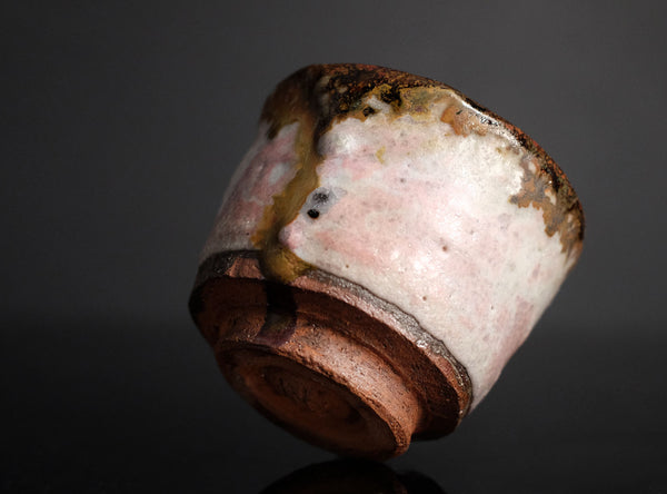 A handmade ceramic sake cup with a textured, organic glaze in pink, brown, and white, featuring a dripping amber glaze detail, set against a dark gray reflective background.