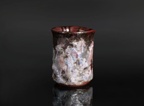 A handmade ceramic cup with a textured, organic surface featuring a mix of white, pink, gray, and dark brown glazes, set against a dark reflective background.