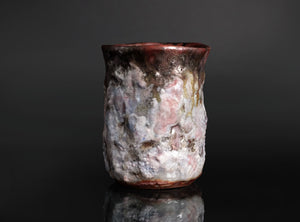 A handmade ceramic cup with a textured, organic surface featuring a mix of white, pink, gray, and dark brown glazes, set against a dark reflective background.