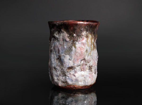 A handmade ceramic cup with a textured, organic surface featuring a mix of white, pink, gray, and dark brown glazes, set against a dark reflective background.