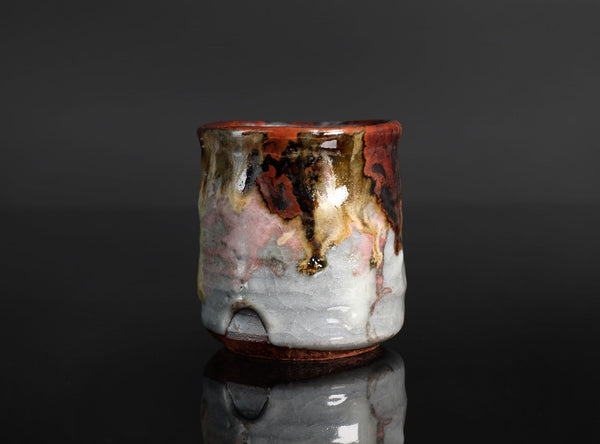 A handmade ceramic cup with a dripping glaze finish, featuring a mix of brown, gold, pink, and gray colors, set against a dark reflective background.