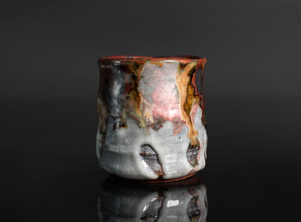 A handmade ceramic cup with a dripping glaze finish, featuring a mix of brown, gold, pink, and gray colors, set against a dark reflective background.