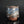 Load image into Gallery viewer, A handmade ceramic cup with a rustic, textured glaze featuring swirls of blue, brown, and white, sitting on a reflective black surface against a dark gray background.
