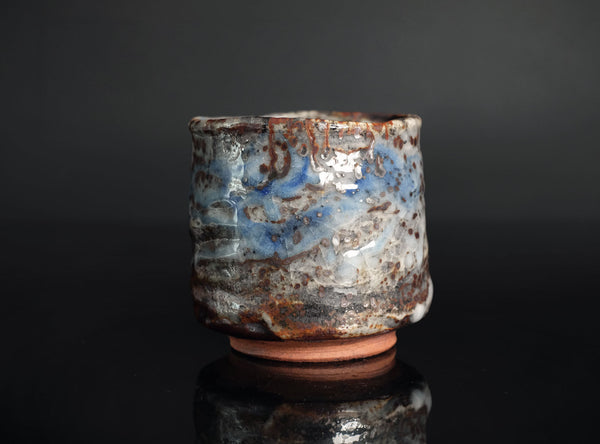 A handmade ceramic cup with a rustic, textured glaze featuring swirls of blue, brown, and white, sitting on a reflective black surface against a dark gray background.