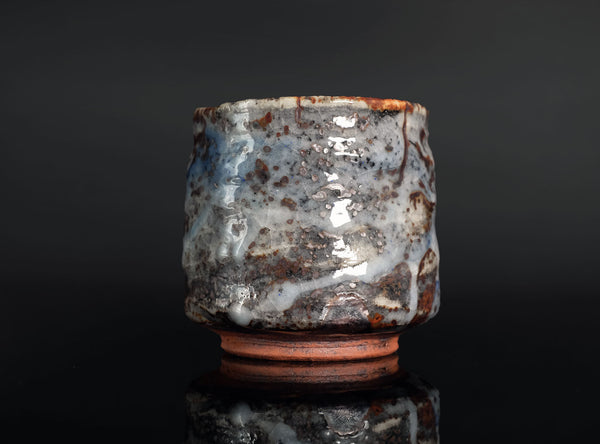 A handmade ceramic tea cup with a textured, organic shape and a glossy glaze featuring mottled gray, white, and rust-brown patterns, set against a dark reflective background.