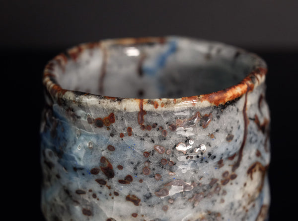 Close-up of a handmade ceramic cup with a textured, organic shape, featuring a glossy glaze of white, blue, and gray with brown and black speckles and drips on a black background.