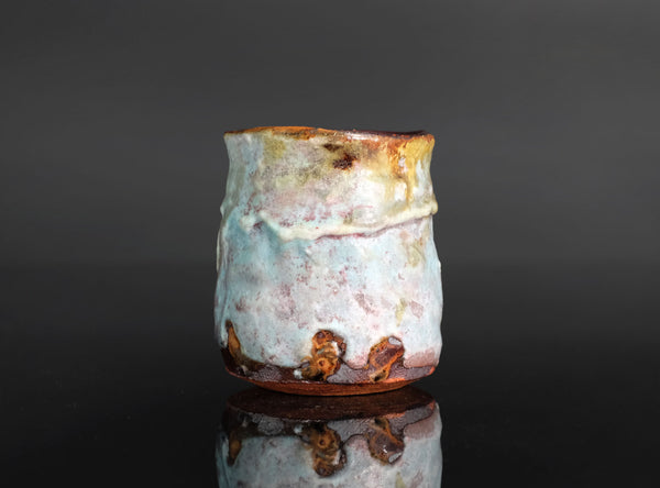 A handmade ceramic cup with a textured, organic shape and a multicolor glaze featuring light blue, white, and earthy brown tones, set against a dark reflective background.