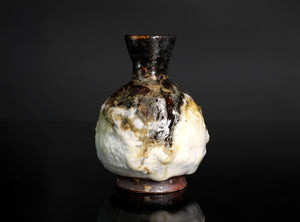 A small ceramic bottle or vase with a bulbous rounded body, dark brown glazed neck, and a mottled white and brown glaze, photographed against a dark studio background.