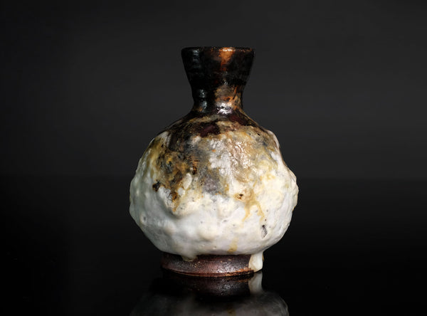 A small ceramic bottle or vase with a bulbous rounded body, dark brown glazed neck, and a mottled white and brown glaze, photographed against a dark studio background.