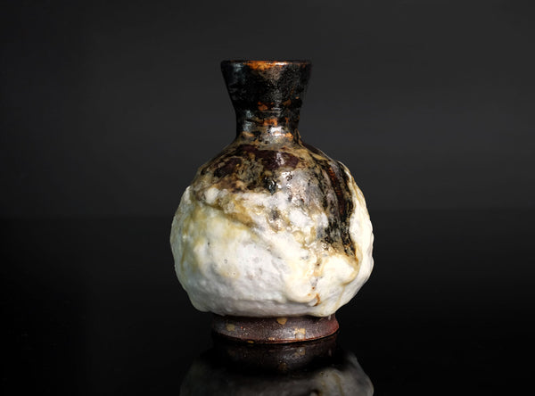 A small ceramic bottle or vase with a bulbous rounded body, dark brown glazed neck, and a mottled white and brown glaze, photographed against a dark studio background.