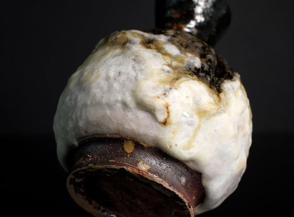 A close-up of a ceramic vase with a rounded, bulbous body covered in thick, textured off-white glaze, dark brown base, and glossy black handle, set against a solid black background.