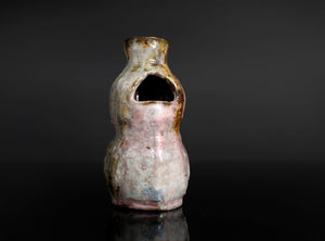 A small handmade ceramic vase with a unique organic shape, featuring a pink and white glaze with brown and gold accents, set against a dark gray background with a reflective black surface.