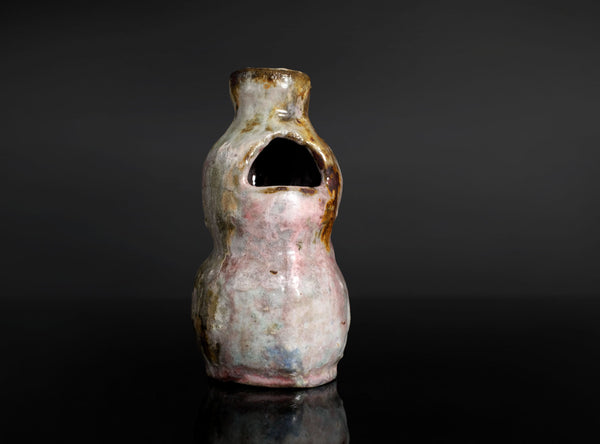A small handmade ceramic vase with a unique organic shape, featuring a pink and white glaze with brown and gold accents, set against a dark gray background with a reflective black surface.