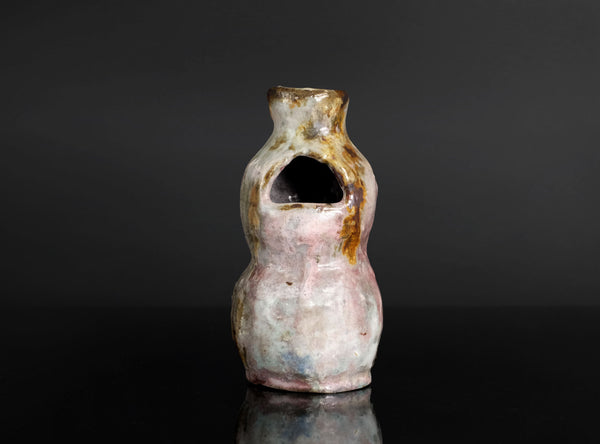 A small handmade ceramic vase with a unique organic shape, featuring a pink and white glaze with brown and gold accents, set against a dark gray background with a reflective black surface.