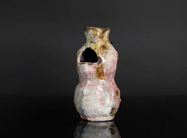 A small handmade ceramic vase with a unique organic shape, featuring a pink and white glaze with brown and gold accents, set against a dark gray background with a reflective black surface.