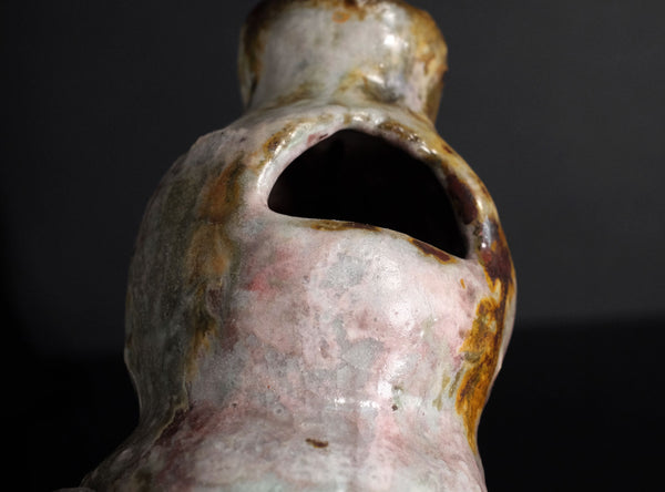 Close-up of a small handmade ceramic vase with a unique organic shape, featuring a pink and white glaze with brown and gold accents, set against a dark gray background with a reflective black surface.