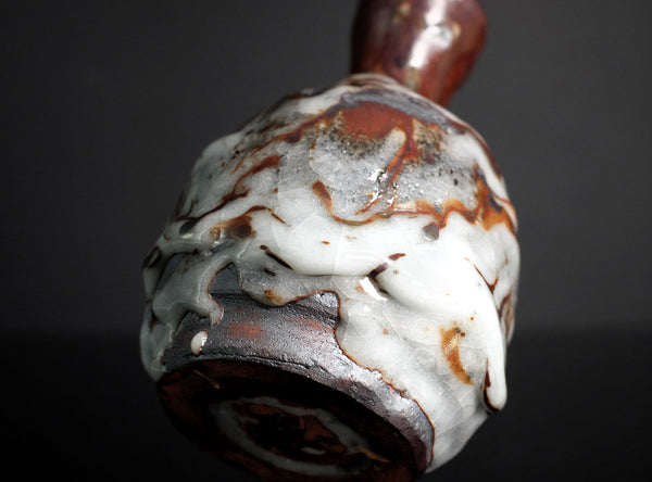 A small ceramic bottle with a rounded body, narrow neck, and flared rim, featuring a glossy glaze with marbled white, brown, and reddish tones, set against a dark reflective background.