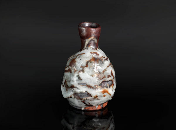 A small ceramic bottle with a rounded body, narrow neck, and flared rim, featuring a glossy glaze with marbled white, brown, and reddish tones, set against a dark reflective background.