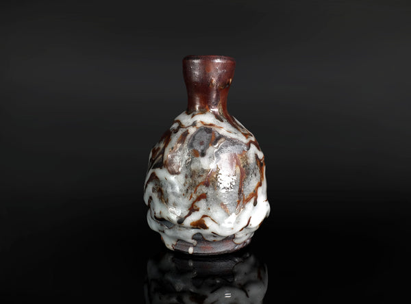 A small ceramic bottle with a rounded body, narrow neck, and flared rim, featuring a glossy glaze with marbled white, brown, and reddish tones, set against a dark reflective background.