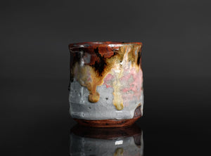 A handmade ceramic cup with a dripping glaze finish, featuring a mix of brown, gold, pink, and gray colors, set against a dark reflective background.