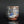 Load image into Gallery viewer, A handmade ceramic cup with a textured, rustic glaze featuring blue, brown, and white tones, standing on a dark reflective surface against a dark gray background.
