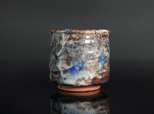 A handmade ceramic cup with a textured, rustic glaze featuring blue, brown, and white tones, standing on a dark reflective surface against a dark gray background.