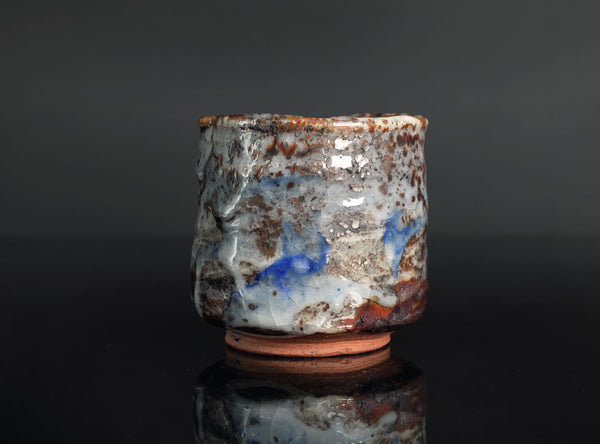 A handmade ceramic cup with a textured, rustic glaze featuring blue, brown, and white tones, standing on a dark reflective surface against a dark gray background.