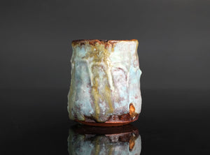 A handmade ceramic cup with a textured, organic shape and a dripping glaze in pale blue, cream, and brown, set against a dark gray background with a soft reflection below.