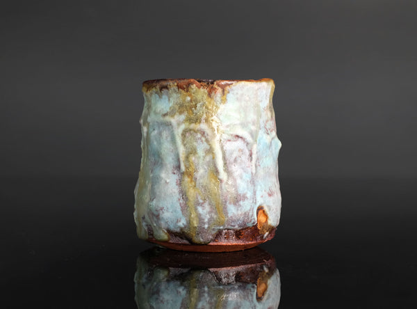 A handmade ceramic cup with a textured, organic shape and a dripping glaze in pale blue, cream, and brown, set against a dark gray background with a soft reflection below.