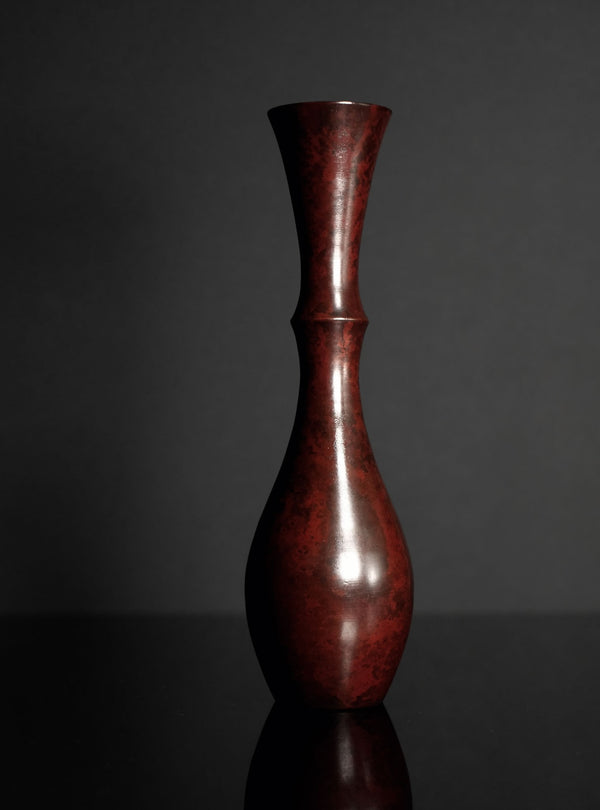 A tall, slender ceramic vase with a flared rim and bulbous base, finished in glossy dark reddish-brown glaze, set against a dark gray background on a reflective black surface.