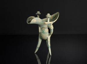 An ancient bronze tri-lobed wine vessel with a patinated greenish-blue surface, standing on a reflective black surface against a dark background.