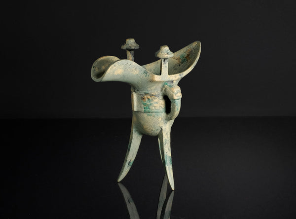 An ancient bronze tri-lobed wine vessel with a patinated greenish-blue surface, standing on a reflective black surface against a dark background.