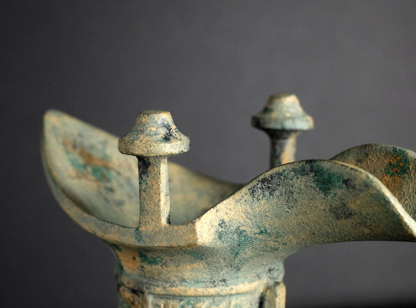 Close-up of an ancient bronze or metal vessel with a weathered green patina, showing the top spouts and curved rim against a dark gray background.