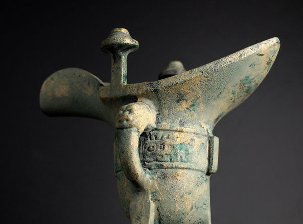 An ancient bronze ritual vessel with a weathered green patina, featuring a curved handle and a decorative emblem on the body, set against a dark background.