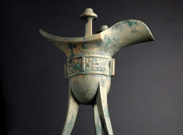 An ancient bronze Chinese wine cup (Jing) with a green verdigris patina, shown against a dark gray background.