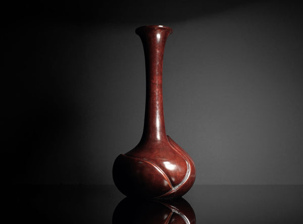 A glossy dark reddish-brown ceramic vase with a bulbous base and a long, slender neck, standing on a reflective black surface against a dark gray background.