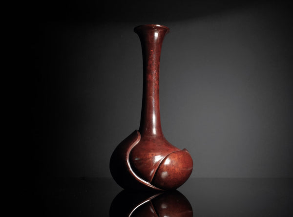 A glossy reddish-brown ceramic vase with a bulbous base and a long, slender neck, displayed on a reflective black surface against a dark gray background.